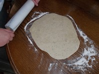 pizza crust being rolled out on a floured wooden table with a white, marble rolling pin