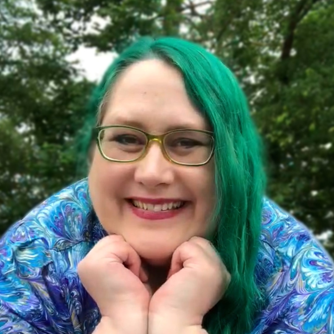 Veronica Bartles, a middle-aged white woman with green hair and green glasses, wearing a shirt with blue, purple, and green swirls reminiscent of peacock feathers, leaning forward with her chin on her hands and smiling in front of a backdrop of trees