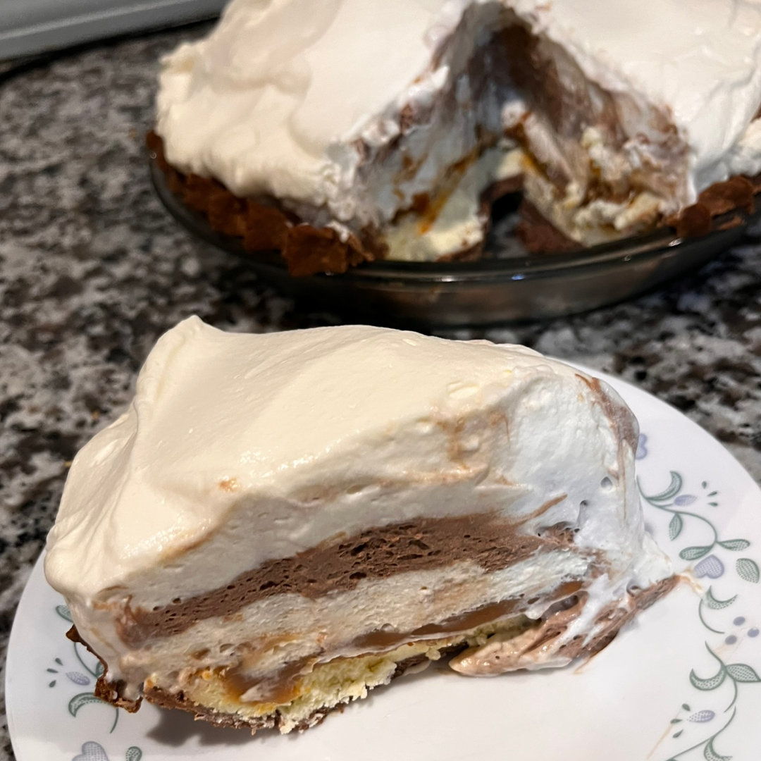 slice of 3.1414926 Ultimate Pie - a 6-layer confection with a chocolate crust, cheesecake base, and layers of caramel, salted caramel mousse, chocolate mousse, and whipped cream - on a white plate with a blue and green floral border, on a granite countertop in front of a full pie, with a single slice removed