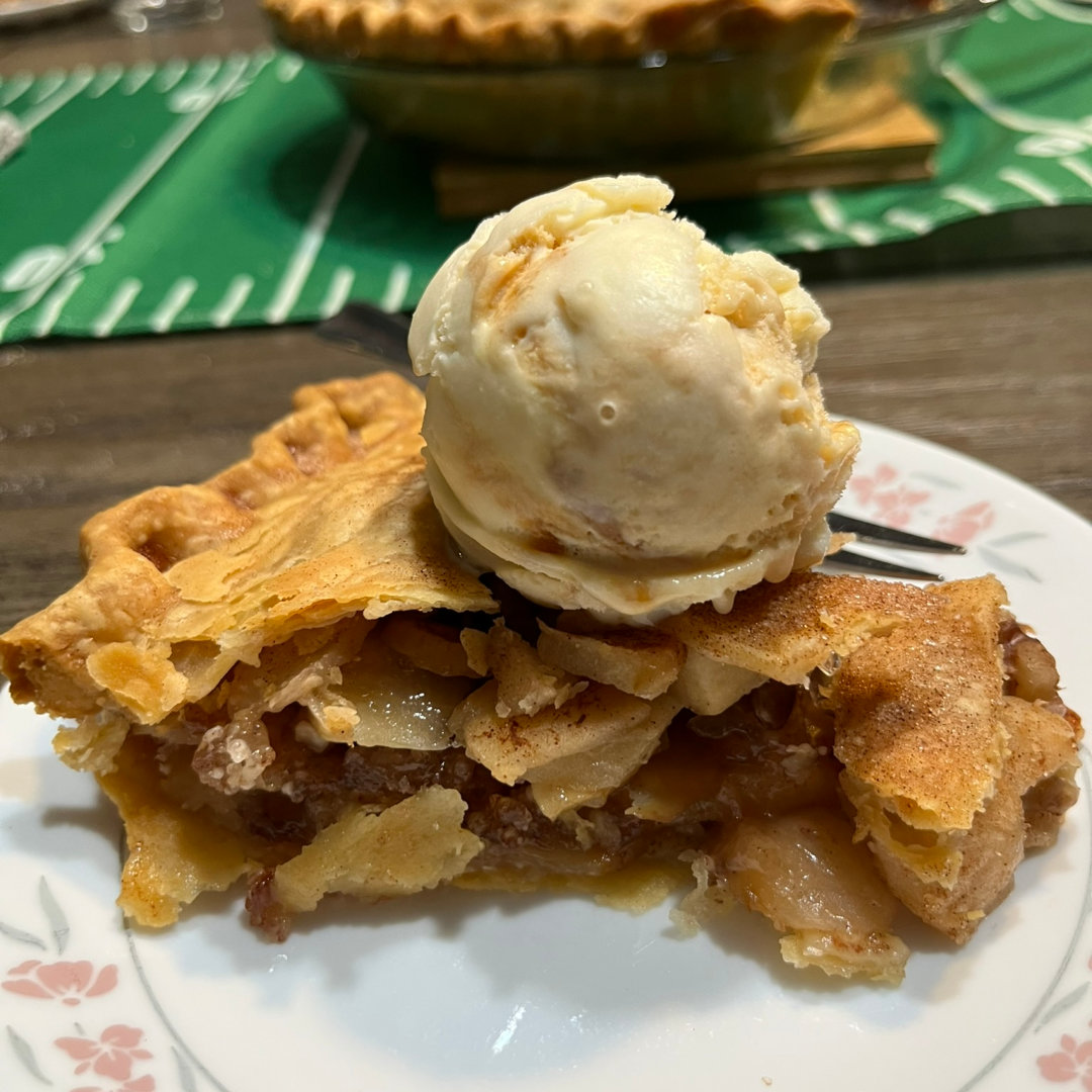 Close-up photo of a slice of Apple Walnut pie on a white plate with a pink and green floral border. The crust is flaky and golden brown, with sprinkles of cinnamon sugar, and there are multiple layers of apple slices and walnuts in the pie. There is a single scoop of slightly-melted caramel ice cream on top of the pie and a fork sitting next to it on the plate. In the background, slightly blurry on a wooden table top behind the pie slice, there is a pie dish with the rest of the pie, sitting in the middle of a green table runner.