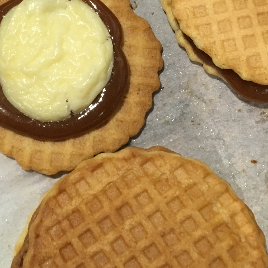 cropped photo of a close-up of Cheesecake Stroopwafel Cookies: 2 thin, vanilla waffle cone wafers with caramel and cheesecake filling sandwiched in between. The cookies are sitting close together on top of a sheet of parchment paper, and the caramel and cheesecake fillings are peeking out from between the wafers of each cookie sandwich. The cookie in the top left corner of the image has the top wafer removed, so you can see the concentric circles of cheesecake, caramel, and cookie wafer.