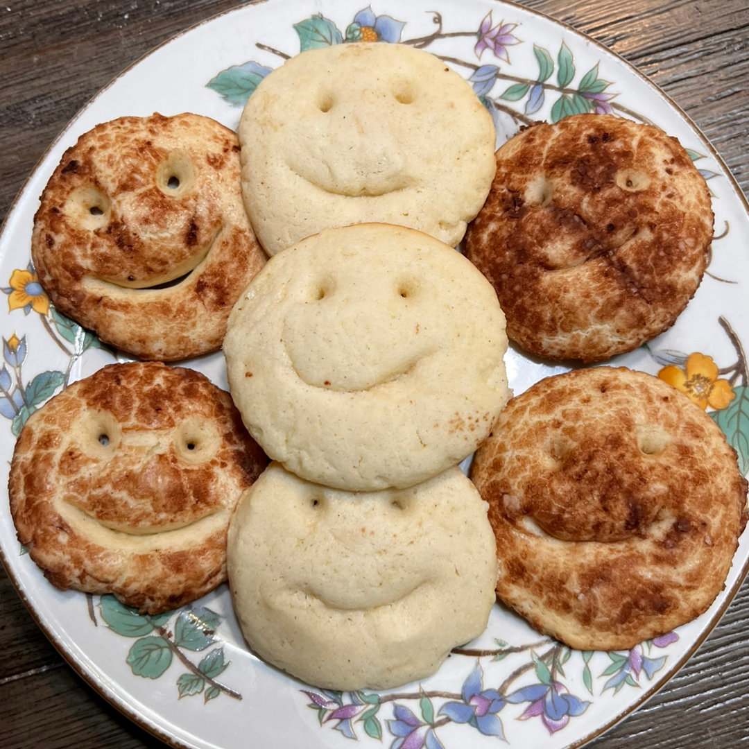Happy Smiles Sugar Cookies - Three rows of sugar cookies with smiley faces cut into them on a white plate with a floral border. The middle row of smiles is unadorned. The rows on the right and left have been rolled in a mixture of cocoa powder and powdered sugar before baking, so they are darker.