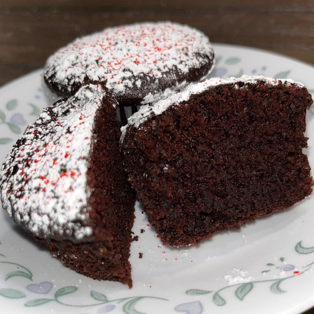 Two dark chocolate cupcakes, dusted with powdered sugar and red sugar sprinkles, sit on a white plate with a green and blue floral border. One cupcake is cut in half, so you can see the detail of the crumb.