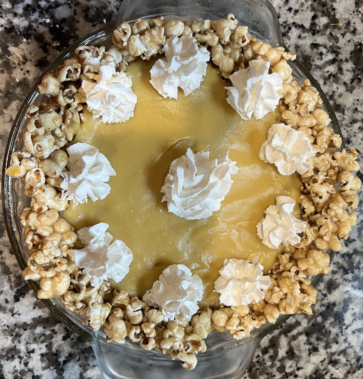 popcorn pie with a caramel corn crust, yellow popcorn custard filling, and whipped cream in a glass pie dish on a mottled granite countertop